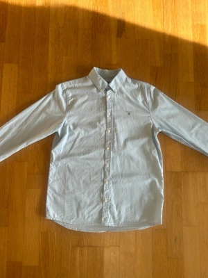 Ljusblå skjorta från Gant - Klassisk ljusblå skjorta från Gant med lång ärm och button-down krage. Skjortan har vita knappar framtill och en liten broderad Gant-logga på bröstet. Perfekt för en stilren och avslappnad look.