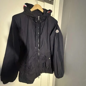 Svart vindjacka från Moncler med huva - Svart vindjacka från Moncler med huva och dragkedja framtill. Jackan har snörning i midjan och på huvan, samt Moncler-logga på ärmen. Huvan har snygga rödvita ränder som ger en sportig vibe. Perfekt för blåsiga dagar och enkel att matcha. (Är öppen för byten) är M men sitter som L