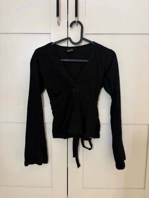 Svart omlottopp med knytband - Svart omlottopp med långa ärmar och knytband i midjan. Djup v-ringning framtill och mjukt, stretchigt material som sitter snyggt på kroppen. 