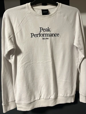 Vit sweatshirt från Peak Performance - Vit sweatshirt från Peak Performance med svart broderad logga och text 'Est. 1986' på bröstet. Tröjan har rund halsringning, långa ärmar och ribbade muddar vid ärmslut och nederkant. Perfekt för en clean och stilren look.