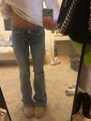 Ljusblå slitna bootcut jeans - Säljer ett par ljusblå bootcut jeans med slitningar och hål över knäna. Jeansen har låg midja och klassisk femficksmodell. Perfekta för en avslappnad och trendig look. Materialet är denim i bomull.