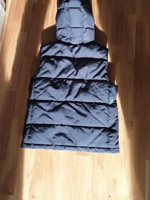 Mörkblå dunväst från Lyle & Scott - Snygg mörkblå dunväst från Lyle & Scott med huva och breda quiltade paneler. Västen har en klassisk, rak passform och är perfekt för lager-på-lager. Märkesdetaljer syns på insidan. Materialet är mjukt och värmande, perfekt för kyliga dagar.