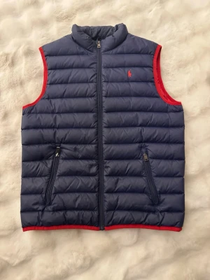 Mörkblå dunväst från Ralph Lauren - Snygg mörkblå dunväst från Ralph Lauren med röda detaljer och dragkedja framtill. Västen har två fickor med dragkedja och klassisk broderad logga på bröstet. Perfekt för lager-på-lager och riktigt chill stil.