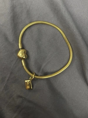 Guldigt armband med hjärtlås och berlock - Snyggt armband i guldfärgad metall med ett hjärtformat lås och en berlock i form av ett kuvert. Armbandet har en stilren, ormliknande kedja och detaljer som ger en trendig look. Perfekt accessoar för att lyfta din outfit. Armband nypris 1200kr berlock nypris 600kr