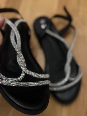 Svarta sandaler med glittriga remmar - Säljer ett par svarta sandaler med tunna, glittriga remmar i silver som korsar sig snyggt över foten. Sandalerna har öppen tå och justerbar rem runt hälen. I form av platta klackar