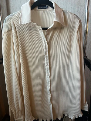 Plisserad vit skjorta från PrettyLittleThing - Beige skjorta med plisserat tyg från PrettyLittleThing. Skjortan har klassisk krage, knappar framtill och långa ärmar. 