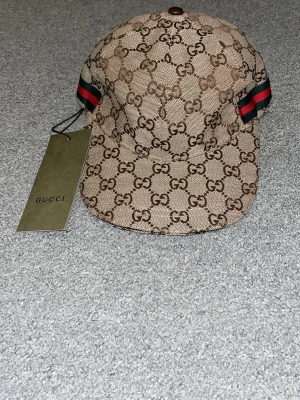 Gucci beige keps med GG-mönster - Säljer en beige keps från Gucci med det klassiska GG-monogrammet över hela kepsen. På sidorna finns gröna och röda ränder som ger en ikonisk touch. Kepsen är tillverkad i canvasmaterial och har en böjd skärm. Perfekt för dig som vill ha en lyxig och trendig accessoar.
