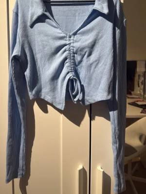 Ljusblå croppad ribbad topp - Trendig ljusblå croppad topp med långa ärmar och krage. Toppen har ribbad struktur och snygg snörning framtill som ger en cool detalj. Perfekt för dig som gillar en modern och avslappnad stil.