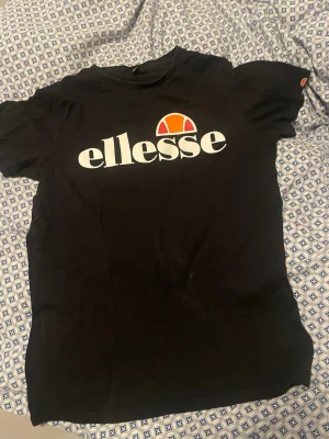 Svart Ellesse t-shirt med logga - Snygg svart t-shirt från Ellesse med stor vit logga och orange detalj framtill. Klassisk rund hals och korta ärmar. Perfekt för dig som gillar sportiga och stilrena plagg. Tillverkad i mjuk bomull för skön känsla hela dagen.