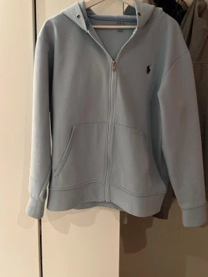 Ljusblå hoodie från Polo Ralph Lauren - Snygg ljusblå hoodie från Polo Ralph Lauren med dragkedja framtill och två stora fickor. Klassisk huva och den ikoniska lilla loggan broderad på bröstet. Perfekt för en avslappnad och stilren look. Fråga något om ni undrar, pris går att diskuteras