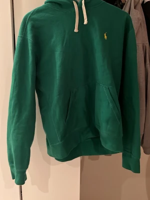 Ralph Lauren Hoodie grön - Snygg grön hoodie från Polo Ralph Lauren med klassisk gul logga broderad på bröstet. Hoodien har vit dragsko i huvan och en stor magficka framtill. Tillverkad i mjukt bomullsmaterial och har ribbade muddar vid ärmslut och nederkant. Pris kan diskuteras