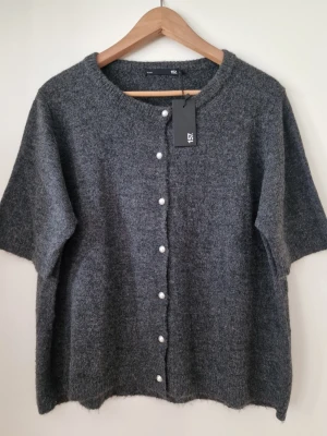 Mörkgrå kofta med pärlknappar Lager 157 - Mysig mörkgrå kofta från Lager 157 med korta ärmar och rund halsringning. Koftan har dekorativa pärlknappar framtill och är stickad i ett mjukt material som ger en avslappnad look. Perfekt att slänga över en t-shirt för extra stil. Storlek L