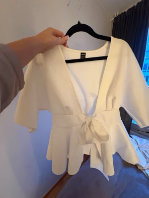 Vit omlottblus med knytband - Stilren vit omlottblus med breda knytband i midjan och v-ringad hals. Blusen har korta ärmar och ett lätt utsvängt peplumliv. Perfekt att matcha med jeans eller kjol för en trendig look.