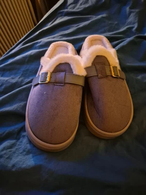 Gråa tofflor med fuskpäls och spänne - Mysiga bruna tofflor med mjukt fuskpälsfoder och dekorativt spänne i guld. Ovandelen är i mockaimitation och sulan är beige. Perfekta för att hålla fötterna varma hemma under kalla dagar.