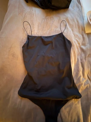 Svart body med tunna axelband - Snygg svart body med tunna axelband och slät, glansig yta. Bodyn har en enkel, stilren design och knäppning i grenen. Aldrig använts så den är helt ny skick, troligen polyester eller liknande.