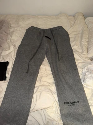 Grå Essentials mjukisbyxor Fear of God - Grå mjukisbyxor från Essentials Fear of God med svart logga på benet och patch framtill. Byxorna har dragsko i midjan och är gjorda i mjukt bomullsmaterial. Perfekta för en chill och avslappnad stil.