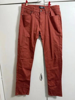 Rostbruna chinos från Dressmann  - Snygga rostbruna chinos från Dressmann med klassisk femficksmodell och raka ben. Tillverkade i mjukt bomullstyg med knapp- och dragkedjegylf. 