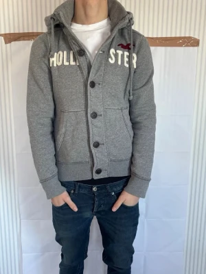 Grå hoodie från Hollister med knappar - Snygg grå hoodie från Hollister med knappar framtill och vit broderad text på bröstet. Har huva med dragsko och två stora fickor. Mjuk bomullsblandning och ribbade muddar vid ärmslut och midja. Perfekt för en chill och avslappnad stil.