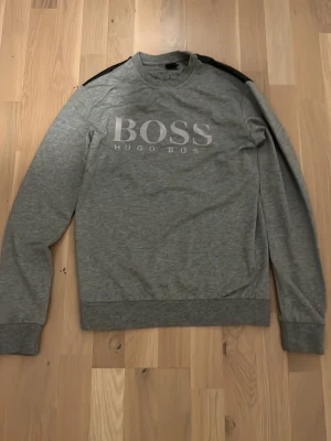 Grå sweatshirt från Hugo Boss - Snygg grå sweatshirt från Hugo Boss med stort BOSS-tryck på bröstet. Tröjan har svarta detaljer på axlarna, rund halsringning och ribbade muddar vid ärmslut och nederkant. Perfekt för en avslappnad och stilren look.