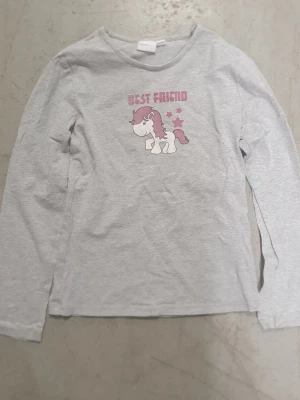 Tröja - Grå långärmad tröja i bomull med tryck av en söt enhörning och texten 'Best Friend' i rosa framtill. Tröjan har en klassisk rund halsringning och raka ärmar. Perfekt för dig som gillar gulliga motiv och mjuka material. Har små fläckar.