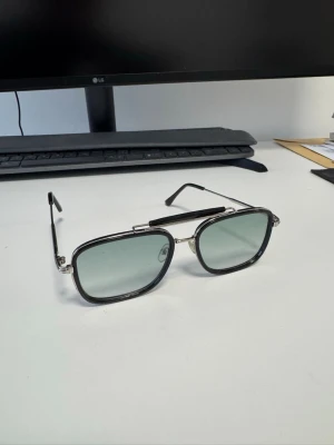 Svarta gröna rektangulära solglasögon - Stilrena solglasögon med rektangulära svarta bågar och silverfärgade detaljer. Glasen är ljusgrå och ger en cool look. Metallbågarna har en modern design som passar perfekt för dig som vill sticka ut.