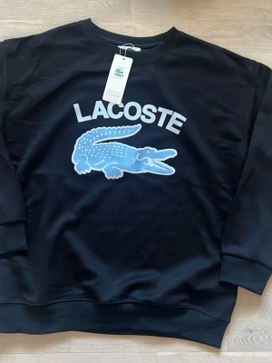 Svart sweatshirt från Lacoste M - Ny och kommer med påse!!💯💯 Cool svart sweatshirt från Lacoste med stort blått krokodilmotiv och vit text på bröstet. Tröjan har rund halsringning och är tillverkad i ett bekvämt och mjukt material, perfekt för en avslappnad streetstyle-look. Pris kan prutas👍👍