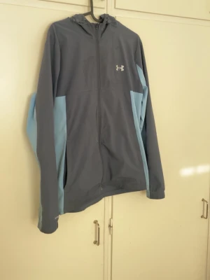 Under Armour gråblå vindjacka - Snygg vindjacka från Under Armour i grått med ljusblå paneler på sidorna och ärmarna. Jackan har huva, hel dragkedja och logga på bröstet. Perfekt för blåsiga dagar och sportiga outfits. Lite smutsig men kan tvättas 