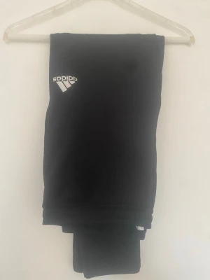 Svarta mjukisbyxor från Adidas - Svarta mjukisbyxor från Adidas med klassisk vit logga på ena benet. Byxorna har resår i midjan och är tillverkade i mjukt material som känns skönt mot huden. Perfekta för chill eller träning, med enkel och stilren design.