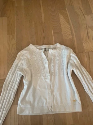 Vit kofta från Hampton Republic - Säljer en vit kofta från Hampton Republic med snyggt kabelstickat mönster och rund hals. Koftan har långa ärmar, knäppning framtill och en liten gulddetalj vid nederkanten. Perfekt att slänga över en topp för en clean look. Passar XS ungefär men är storlek 158/164.