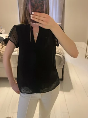 Svart transparent blus från Vila - Snygg svart blus från Vila med korta, transparenta ärmar i mesh och prickigt mönster. Blusen har knappar framtill och en lätt v-ringning. Perfekt att styla med både jeans och kjol för en trendig look.