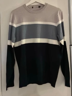 Randig långärmad tröja i blått och grått - Snygg långärmad tröja med breda horisontella ränder i ljusgrått, mörkblått, vitt och blågrått. Tröjan har rund halsringning och en stilren, avslappnad passform. Perfekt för en trendig och enkel look.