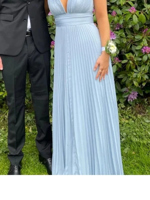 Ljusblå plisserad långklänning - Snygg ljusblå långklänning med djup v-ringning och breda axelband. Klänningen har plisserad kjol, omlottliv och knytband i midjan som man kan knyta både fram och bak beroende på hur man öskar. Perfekt till bal eller annan festlighet i sommar