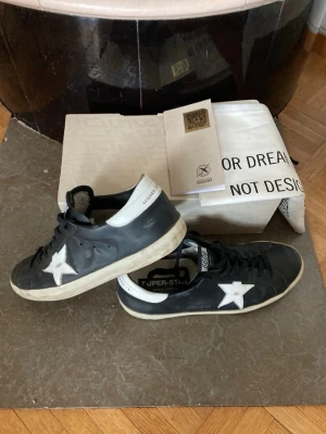 Golden Goose svarta sneakers med stjärna - Svarta sneakers från Golden Goose med vit stjärndetalj på sidan och vit hälkappa. Klassisk låg modell med slitna detaljer och svart-vit sula. Skorna har snörning och märkets logga på plösen. Perfekta för en cool och avslappnad streetstil. Följdesedel med kommer. Pris går att diskutera!!