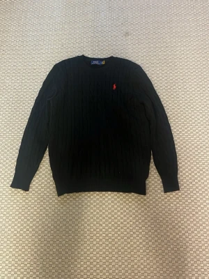 Ralph Lauren Tröja - Kabelstickad svart tröja från Polo Ralph Lauren med rund halsringning och den klassiska röda logotypen broderad på bröstet. Långa ärmar och ribbade muddar vid ärmslut och nederkant. Perfekt för en stilren och tidlös look.