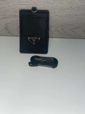 Svart korthållare från Prada - Snygg svart korthållare från Prada i strukturerat läder med silverfärgad logga framtill. Kommer med en matchande svart rem som kan fästas i metallringen upptill. Perfekt för att hålla koll på kort och ID med stil.