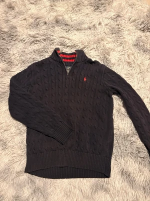 Mörkblå stickad tröja från Ralph Lauren - Mörkblå stickad tröja från Ralph Lauren med halv dragkedja och röd broderad logga på bröstet. Tröjan har kabelstickat mönster, ribbade muddar och krage med röd- och grårandigt foder. Perfekt för en snygg och avslappnad stil.