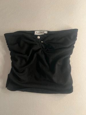 Svart bandeau tubtopp från Kappahl - Svart bandeau tubtopp från Kappahl med snygg rynkning framtill och dekorativa pärldetaljer. Toppen är ärmlös och har en lätt strukturerad yta som ger en cool känsla. Perfekt att styla med jeans eller kjol för en trendig look.