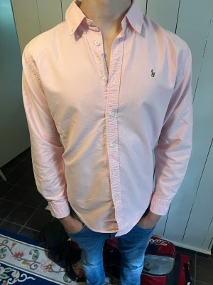 Rosa Ralph lauren skjorta - Säljer en klassisk ljusrosa skjorta från Ralph Lauren med button-down krage och broderad logga på bröstet. Skjortan har långa ärmar, knappar framtill och är tillverkad i bomull. Perfekt för en stilren och avslappnad look. Passar ca S-M