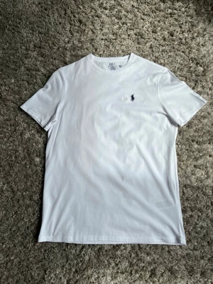 Vit t-shirt från Polo Ralph Lauren - Klassisk vit t-shirt från Polo Ralph Lauren med liten broderad logga på bröstet. T-shirten har rund halsringning och korta ärmar, tillverkad i mjuk bomull för en skön känsla. Perfekt basplagg till garderoben.