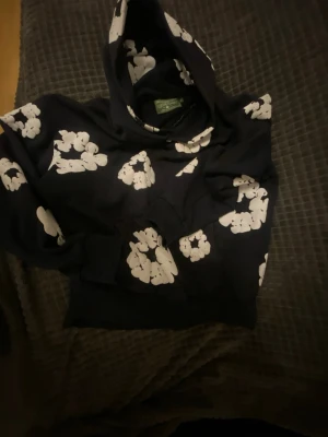 Denim tears hoddie - Snygg svart hoodie med stora vita blommönster över hela tröjan. Hoodien har en klassisk huva och ribbade muddar vid ärmslut och nederkant. Perfekt för dig som gillar en unik och trendig stil.