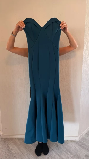 Blå långklänning med hjärtformad urringning - Elegant blå långklänning med hjärtformad urringning och figurnära passform upptill. Klänningen har en utsvängd nederdel och är axelbandslös, vilket ger en stilren och modern look. Perfekt för speciella tillfällen när du vill sticka ut.