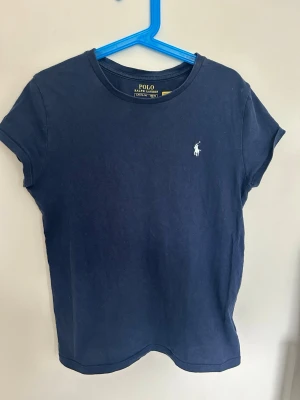 Marinblå t-shirt från Polo Ralph Lauren - Snygg marinblå t-shirt från Polo Ralph Lauren med klassisk vit logga på bröstet. T-shirten har rund halsringning och korta ärmar. Perfekt basic-plagg i mjuk bomull som passar till allt.