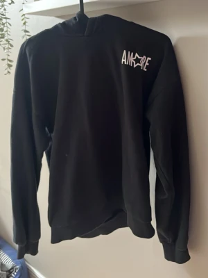 Svart hoodie med AMORE-tryck - Snygg svart hoodie med vitt AMORE-tryck på bröstet och en stjärndetalj i loggan. Tröjan har rund halsringning, långa ärmar och ribbade muddar. Perfekt för en avslappnad och trendig stil.