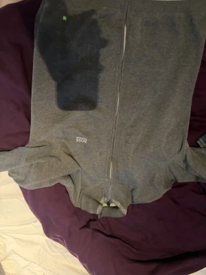 Grå ziptröja från Hugo Boss - Snygg grå ziptröja från Hugo Boss med diskret logga på bröstet och gröna detaljer vid dragkedjan. Tröjan har ribbade muddar och är tillverkad i mjukt material som känns skönt mot huden. Perfekt för en stilren och avslappnad look.