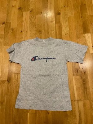 Grå Champion t-shirt med logga stl S - Snygg grå t-shirt från Champion med klassisk logga i blått och rött på bröstet. T-shirten har rund hals och korta ärmar, perfekt för en avslappnad och sportig stil. Tillverkad i mjuk bomull för skön komfort.