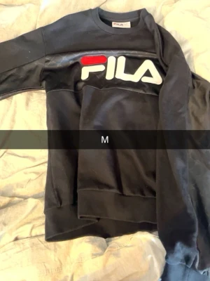 Svart sweatshirt från FILA - Säljer en svart sweatshirt från FILA med stor vit och röd logga framtill. Tröjan har rund halsringning, långa ärmar och ribbade muddar. Materialet är mjukt och bekvämt, perfekt för en avslappnad stil.