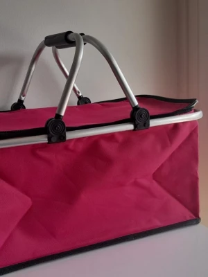 Rosa shoppingväska med aluminiumhandtag - Snygg och rymlig shoppingväska i stark rosa färg med rektangulär form. Väskan har dubbla aluminiumhandtag med svarta plastdetaljer och svart kantband. Perfekt för att bära hem mat eller andra grejer från stan.