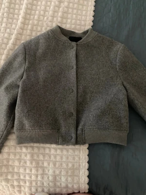 Grå bomberjacka i ullmix - Snygg grå bomberjacka med minimalistisk design och ribbade muddar. Jackan har rundad krage och knäppning framtill med dolda tryckknappar. Perfekt för dig som gillar stilren och enkel look.