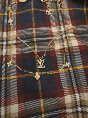 Louis Vuitton guldhalsband med pärlor - Dubbelkedjat halsband i guldfärgad metall från Louis Vuitton med LV-logga och små blommor dekorerade med vita pärlor. Kedjan har fina detaljer och hängsmycken som ger en lyxig känsla. Perfekt accessoar för att lyfta din outfit.