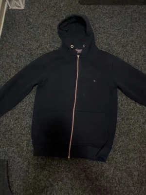 Svart hoodie med dragkedja Tommy Hilfiger - Svart hoodie från Tommy Hilfiger med dragkedja framtill och klassisk huva med metallöljetter. Liten broderad logga på bröstet och två stora fickor framtill. Perfekt för en chill och stilren look.
